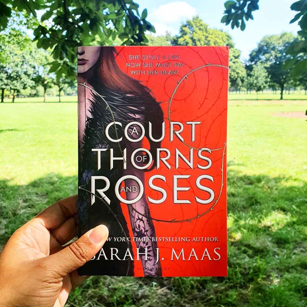 A copy of 'A Court of Thorns and Roses' is being held in front of trees and grass in the background. It's a sunny day. 