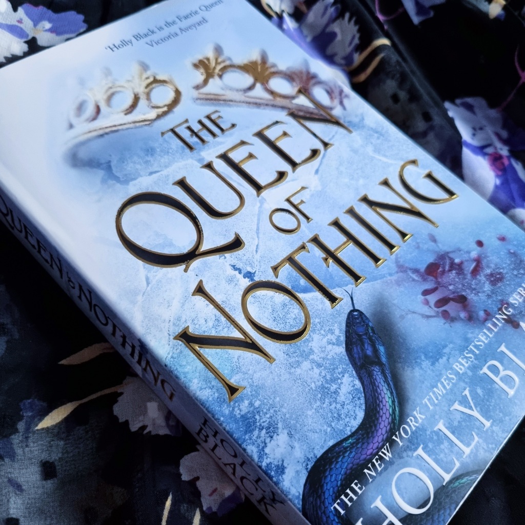 a close up of a paperback copy of 'the queen of nothing.' it is on top of a purple and black floral blouse. 
