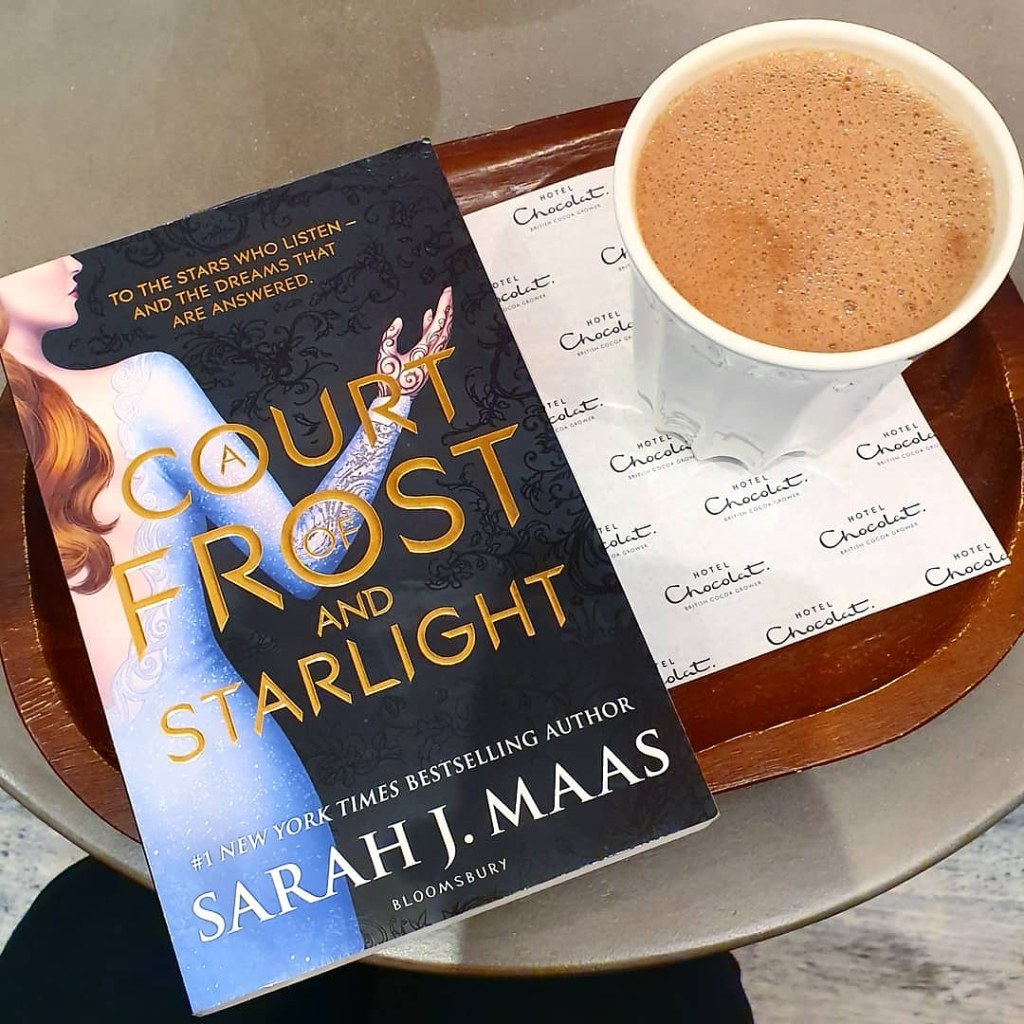 A paperback copy of A Court of Frost and Starlight next to a cup of hot chocolate from Hotel Chocolat. 